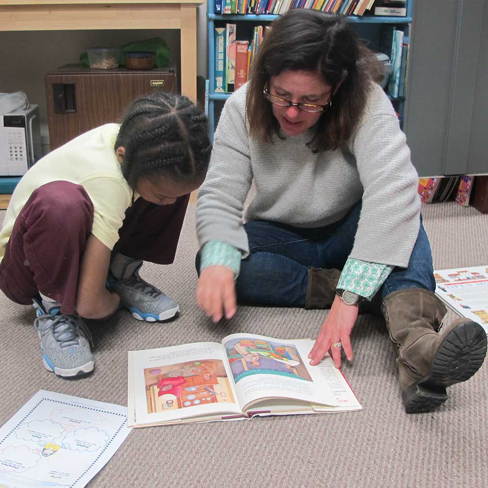 Brown Memorial tutor sits on floor reading a book with a student.